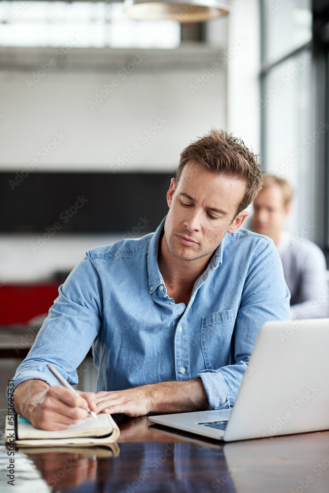 Poster staying connected to his creativity. a man writing notes while working on a laptop in an office.