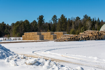 stacks  of milled wood and raw logs outside of limber mill in winter.