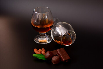 Two glasses with cognac on a black background, next to chocolates, mint leaves and almonds.