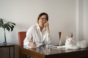 Serious middle-aged business woman wearing eyeglasses, white linen shirt with long straight hair working at home office with laptop, white cat lying on table.Distance work and pet concept.Copy space.