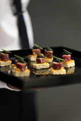 The waiter holds a Set of festive appetizer canapes on bread on a black tray as an event dish. Buffet table. Catering. Delicacy. Haute cuisine. 