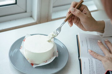 Happy asian woman wearing white dress sitting and eating cake on wooden plate in dessert cafe while reading a book