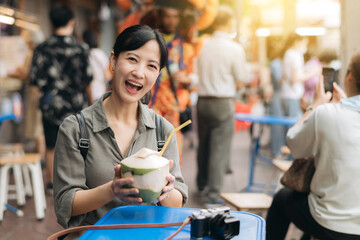 Happy young Asian woman backpack traveler drinking a coconut juice at China town street food market in Bangkok, Thailand. Traveler checking out side streets.