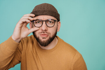 Young beardy caucasian guy in glasses and hat looks at camera with puzzled face expression. Pensive...