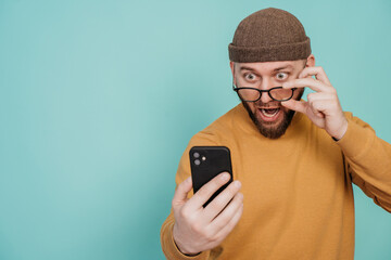 Amazed young man in hat and sweater holds phone takes off his spectacles in discouraged face...
