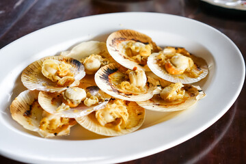 Grilled scallops in the shell with butter, a fresh seafood menu of a restaurant, served on a white plate on the wooden table with Thai seafood sauce.