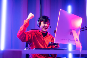 Professional gamer casting podcast walkthrough review. Young asian man sitting on chair with computer pc and microphone. Happy male Streamer wearing headphone playing game online in room neon light © Chanakon