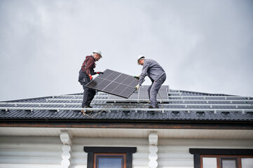 Mounters building solar panel system on roof of house. Men workers in helmets carrying photovoltaic...