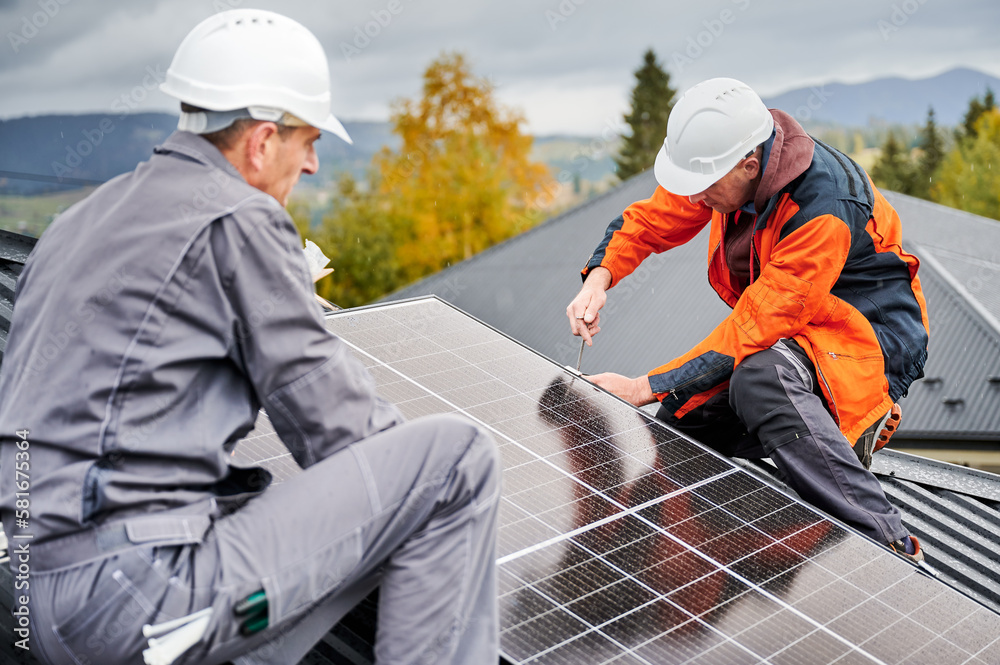 Wall mural Technicians installing photovoltaic solar panels on roof of house. Men engineers in helmets building solar module system with help of hex key. Concept of alternative, renewable energy.