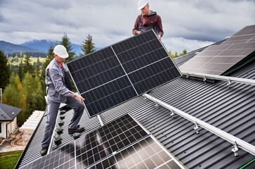 Engineers building solar panel system on roof of house. Men workers in helmets carrying photovoltaic solar module outdoors. Concept of alternative and renewable energy. - obrazy, fototapety, plakaty