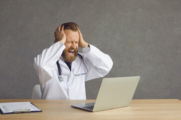 Tired crazy male doctor sitting at office table with computer crying holding hands on head stressed by report deadline, medical bureaucracy workload, work schedule overload or new healthcare reform