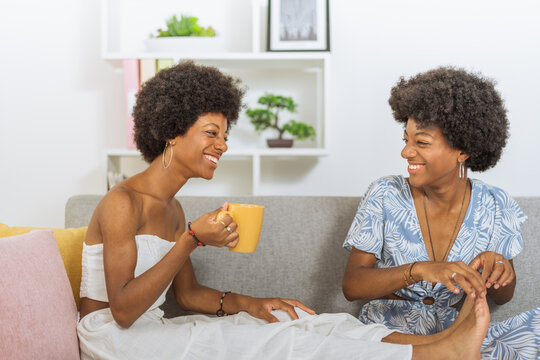 Identical black twin sisters, laughing loudly, one holding the feet of the other who is holding a cup of coffee