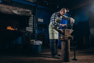 Senior an artisan blacksmith knocks with a hammer on iron to shape against the background of a burning forge