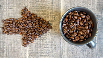 a dark mug filled with whole grains of coffee on the table.  an arrow made of coffee beans points to the cup.  view from above .  cheerfulness, energy