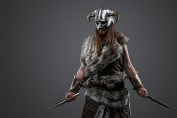 Shot of nordic vandal with fur and helmet holding two daggers against grey background.