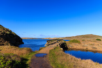 Wooden footbridge over blue water along dirt trail in dry coastal landscape