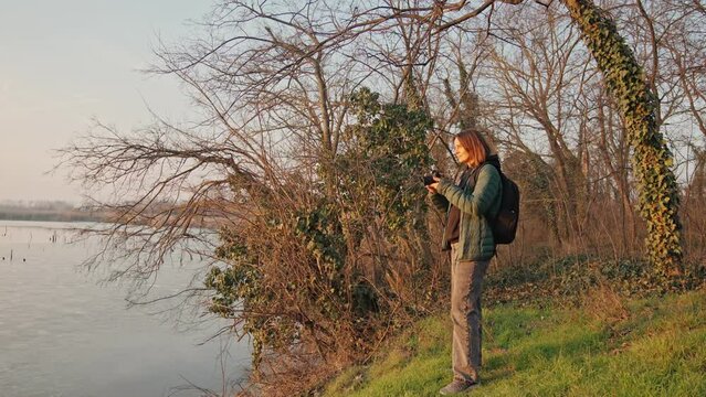 Slow-motion cinematic shot. A woman professional photographer taking a picture of a landscape while standing by the lake at the sunset time