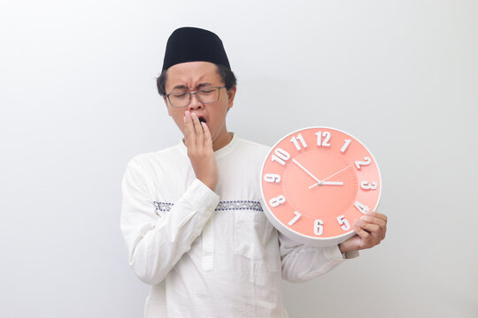 Portrait of young sleepy Asian muslim man yawning with hand covering his mouth while holding a clock that shows 3 am. Isolated image on white background
