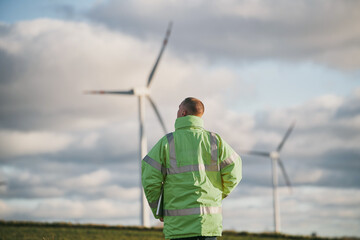 Back view of young male engineer in a wind turbine farm in the background.