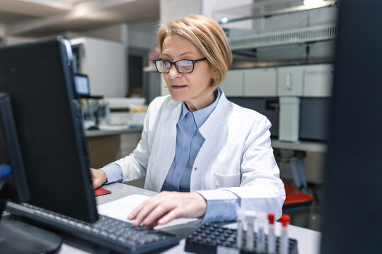 Female Scientists Working on their Computers In Big Modern Laboratory. Various Shelves with Beakers, Chemicals and Different Technical Equipment is Visible.