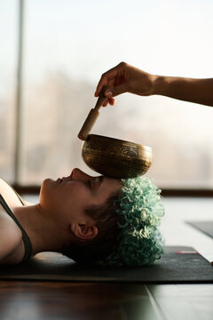 Vertical image of young girl lying on the floor on exercise mat and relaxing with singing bowl on her face during practice