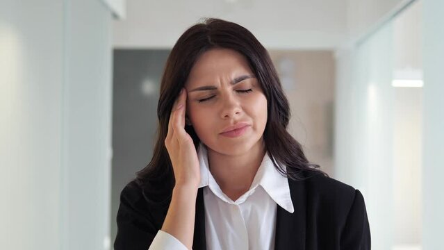 Businesswoman in professional attire is dealing with a painful headache or migraine in office lobby, likely caused by work related stress and lack of self care. Need medical attention. Stress.