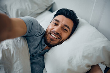 Happy man taking selfie while relaxing in bedroom and looking at camera.