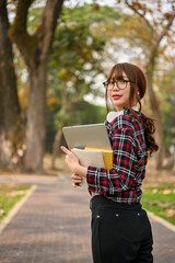 Asian female college student holding her laptop and book, walking along the park.