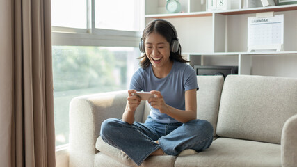 Obraz premium Asian woman playing games on mobile phone and wearing headphones having fun, Play games on the sofa in the living room on weekends, Resting at home, Comfort zone, Touch screen mobile phone.