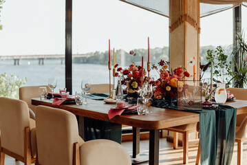 A table in a restaurant on the river bank with panoramic windows, professionally decorated with glass and crystal details, fresh red and orange flowers and candles in candlesticks