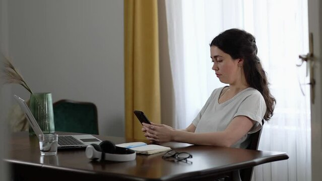 Office work. Young Caucasian woman using smartphone sitting at desktop. Freelance and remote work