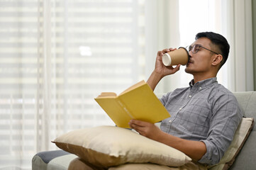 Side view of a smart Asian man sipping coffee and reading book while relaxing in living room.