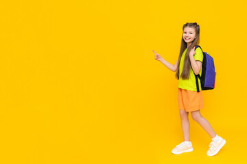 A child with a full-length backpack. A little girl with a satchel in summer shorts and a T-shirt points to an empty copy space. Yellow isolated background. Banner. A place for your text.