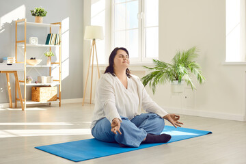 Fototapeta Cheerful attractive overweight young woman doing yoga at home. Relaxed plus size woman sitting on mat with her legs crossed, meditating in lotus position. Healthy lifestyle, relaxation concept obraz