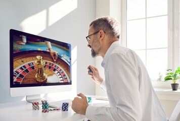 Mature man gambler in glasses and white shirt places bet in online casino gambling game looking at computer screen sitting at table. Gambling, betting, online casino, gambling addiction concept.