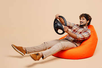 Full body young Indian man wearing brown shirt casual clothes sit in bag chair hold steering wheel...