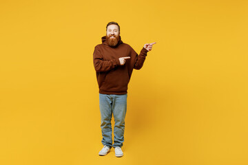 Fototapeta na wymiar Full body young redhead caucasian man wearing brown hoody casual clothes pointing indicate on workspace area copy space mock up isolated on plain yellow background studio portrait. Lifestyle concept.
