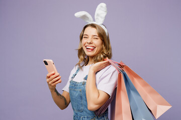 Fototapeta na wymiar Young fun woman wears casual clothes bunny rabbit ears hold shopping paper package bag use mobile cell phone isolated on plain light pastel purple background studio Happy Easter sale buy day concept.