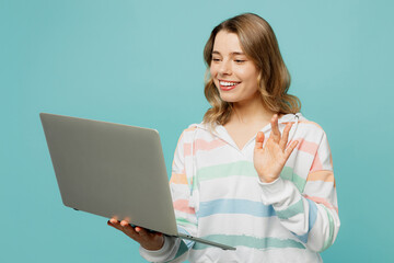 Ycoung IT woman wear striped hoody hold use work on laptop pc computer talking online get video call waving hand isolated on plain pastel light blue cyan background studio portrait. Lifestyle concept.