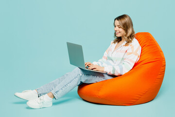 Full body young IT smiling happy woman wear striped hoody sit in bag chair hold use work on laptop...
