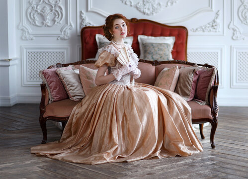Young red haired woman in ball historic dress and fan in hand in vintage room sitting on sofa. Portrait of aristicratic woman in historically decorated room.