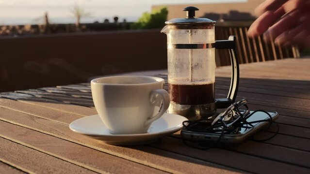 Caf&eacute; caliente humeante de cafetera de &eacute;mbolo sobre una mesa de madera con luz de amanecer en una terraza con vistas al mar y jardinera de fondo, con mano de hombre que sirve caf&eacute;.