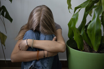 A teenage girl sits with her arms clasped on her knees on the floor against a wall next to houseplants. She's upset and sad. The problem of loneliness, bullying, mental health in the modern world