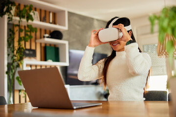 A smiling Asian woman with a VR headset, working on her laptop at home.