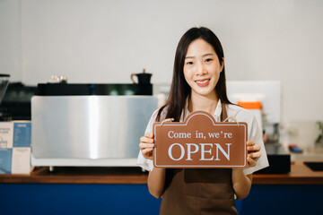 Young female entrepreneur hanging a welcome sign in front of a coffee shop. Beautiful waitress or...