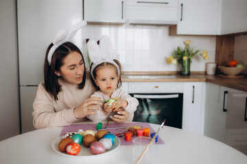 Mother and her daughter painting eggs. Happy family preparing for Easter. Cute little child girl wearing bunny ears.