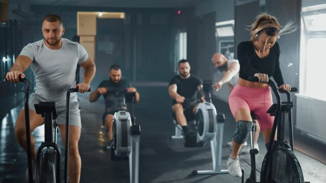 Attractive woman bodybuilder and man doing exercise on stationary bike an two men work out together on rowing gym machines slow. Group training