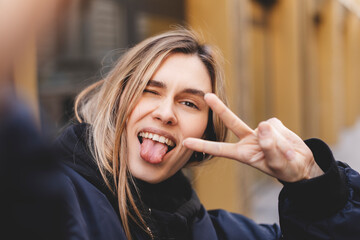 Stylish girl with blonde hair making selfie outside. Cool woman in fashion bomber showing peace...