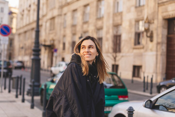 Fototapeta premium Happy blonde woman wear navy blue bomber jacket walking in the street and smiling, she look up. Stylish girl in fashion outfit walking over the city. What a beautiful day out.