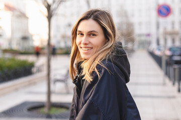 Fototapeta na wymiar Happy blonde woman wear navy blue bomber jacket walking in the street and smiling to camera. Stylish girl in fashion outfit walking over the city. What a beautiful day out. Girl turn around.
